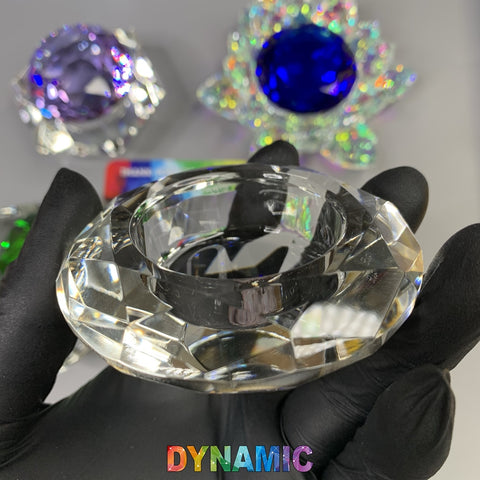 A hand in a black glove holds one of the faceted, oval-clear lead crystals from the CRYSTAL DAPPEN DISH collection by Dynamic Nail Supply. In the background, you can see other crystals from this collection: a blue crystal with holographic petals featuring a lotus shape, alongside purple and green crystals on a gray surface. The word "DYNAMIC" is boldly displayed at the bottom in colorful block letters.