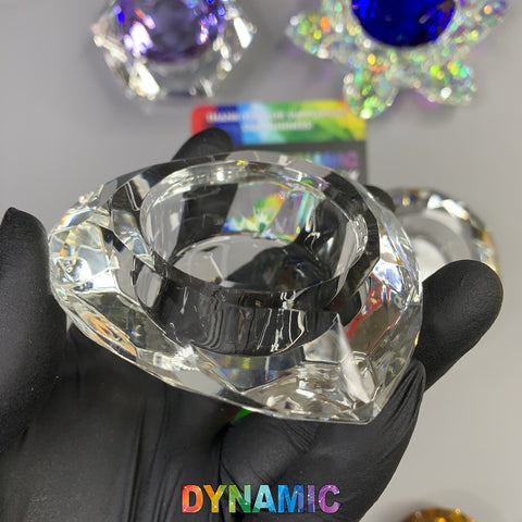 A gloved hand displays a crystal-cut ring stand, its surfaces shimmering as if they were crafted from lead crystal. In the background, a holographic card and a collection of crystalline objects can be seen, including a Dynamic Nail Supply CRYSTAL DAPPEN DISH in lotus shape and a striking blue bowl with petal-like edges. The bottom of the image is adorned with the vibrant word "DYNAMIC.