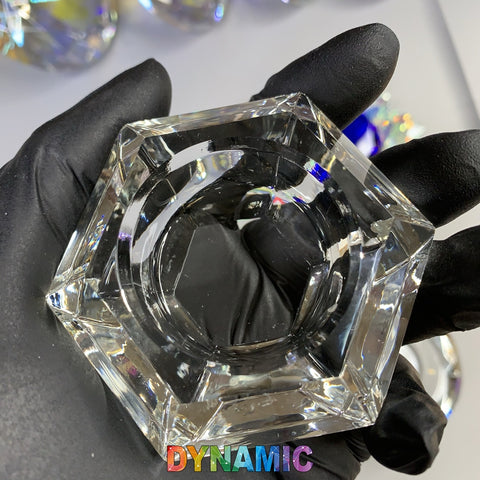 A hand wearing a black glove holds a clear hexagonal glass object reminiscent of a Dappen dish. Below, the brand name "Dynamic Nail Supply" is displayed in colorful, gradient letters. The background features blurred, reflective surfaces, suggesting additional glass objects from the CRYSTAL DAPPEN DISH series made of lead crystal.