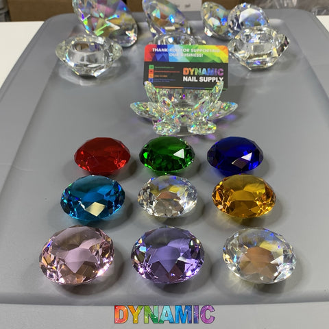 An array of large, vibrant gemstones meticulously placed on a gray surface captivates with hues of red, green, blue, teal, yellow, purple, and pink. A transparent lead crystal lotus from the CRYSTAL DAPPEN DISH collection by Dynamic Nail Supply is accompanied by a branded card to create an elegant display while the word "DYNAMIC" gleams at the bottom.