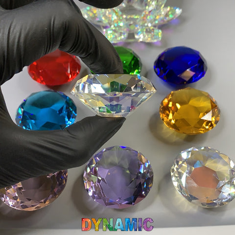 A gloved hand delicately holds a clear, faceted lead crystal gem from the Dynamic Nail Supply collection of CRYSTAL DAPPEN DISH - HEXAGON - ROUND - HEART - LOTUS SHAPE over a white surface. Surrounding it are colorful gems in red, green, blue, orange, yellow, and purple. Each gem is cut with precision, reflecting light. Below, the brand name "Dynamic Nail Supply" is displayed in a rainbow gradient font.