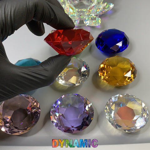 A gloved hand holds a large red gemstone above eight other multicolored gemstones, similar to the exquisite styles of Dynamic Nail Supply's CRYSTAL DAPPEN DISH - HEXAGON - ROUND - HEART - LOTUS SHAPE. Arranged in a grid on a gray surface, the gemstones showcase vibrant colors like blue, yellow, teal, purple, clear, and pink. At the bottom of the image, the word "DYNAMIC" is printed in rainbow hues.