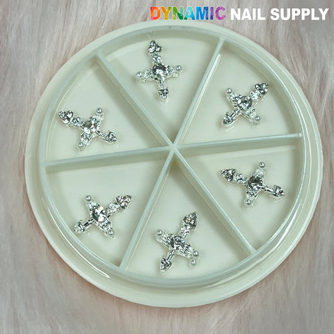 A round, segmented container holds six Silver Cross Charm for Nails Art Designer pieces, each embellished with small gemstones. Positioned on a soft, textured pink surface, this design exemplifies the craftsmanship of Nails Art Designer. The brand name Dynamic Nail Supply is stylishly displayed in the top right corner.