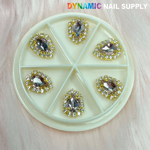 A round container with a clear lid displays six teardrop-shaped nail art gems, each encircled by small rhinestones. Included in this collection are the stunning Rose Gold Heart Rhinestone Nail Charms. These gems are set against a pink fur backdrop, with the brand "Dynamic Nail Supply" featured in the top right corner.