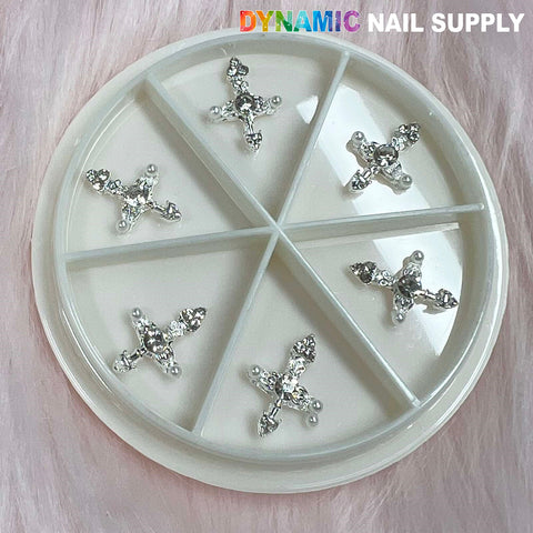 A round container with a clear lid is sectioned into six compartments, each containing a Silver Cross Charm for Nails Art Designer adorned with rhinestones and small pearls. The backdrop features soft, pink furry fabric, while the brand name Dynamic Nail Supply is printed in the top right corner, highlighting their exquisite creations.