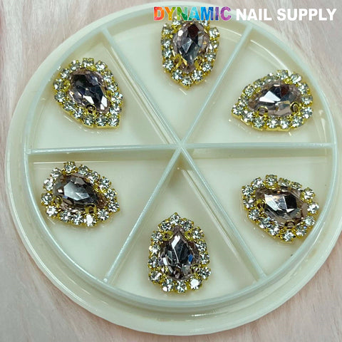 A circular embellishment wheel from Dynamic Nail Supply, featuring six compartments filled with pear-shaped, jeweled nail charms. The large, multifaceted jewels boast a silver center encircled by small gold and clear rhinestones. "Dynamic Nail Supply" is elegantly displayed at the top.