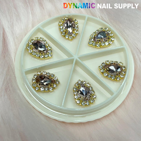A round plastic container featuring six compartments, each filled with teardrop-shaped rhinestones named "Rose Gold Heart Rhinestone Nail Charms." These charms are silver with a gold border adorned with small, clear gems. The container's background is a soft, furry pink, and the brand name "Dynamic Nail Supply" is printed at the top right.