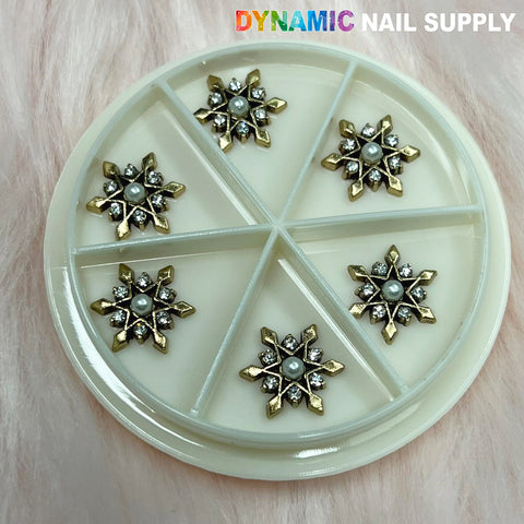 A circular container features six compartments, each containing antique star flower snowflake charms in metallic gold and silver. Each charm is embellished with a central pearl and small rhinestones, set against a soft pink fuzzy background. The "Dynamic Nail Supply" logo adorns the top right corner, making it perfect for any nail art designer.