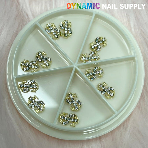 A circular white plastic container featuring six compartments, each containing stunning Gold Dollar Sign Charms for nail art adorned with rhinestones. Placed on a gentle pink background, the container is marked with "Dynamic Nail Supply" in bold multicolored and white lettering.