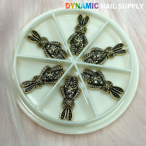 A round, sectioned container showcases six metallic, bunny-shaped nail charms in antique designs, each adorned with intricate details and embedded rhinestones. The backdrop is a soft and fuzzy texture reminiscent of light pink fur. In the top right corner, the brand name Dynamic Nail Supply is elegantly displayed in gradient colors.