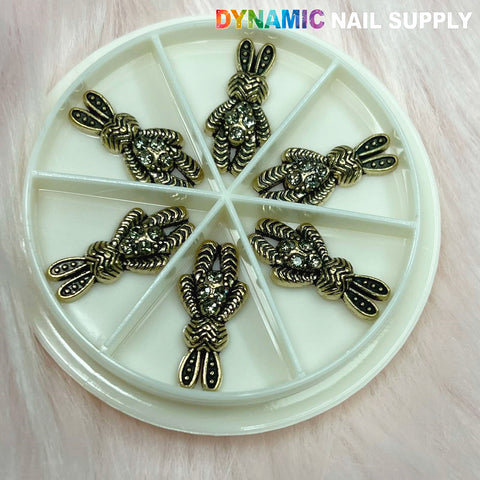 A round white container with six compartments features metal nail charms in the shape of rabbits, showcasing intricate antique-style designs. Against a soft pink backdrop, "Dynamic Nail Supply" is prominently displayed in colorful letters at the top right corner, emphasizing its unique nail art collection. The product is called Antique Bunny Charm for Nails Art Designer.