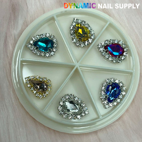 A circular plastic container with six compartments holds heart-shaped, gem-like nail charms. Each is surrounded by small clear rhinestones and features a vibrant centerpiece in shades of blue, yellow, multicolor, silver, and dark blue. The text reads Dynamic Nail Supply.