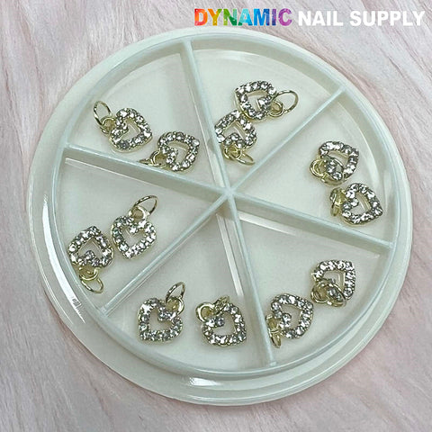 A round plastic container, featuring compartments, contains multiple heart-shaped dangles with rhinestones from the Heart Shaped Dangles Nail Charms set. The name Dynamic Nail Supply is displayed at the top right, highlighting its dedication to quality. The container is placed on a light-colored, textured surface.