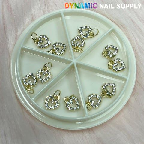 The Heart Shaped Dangles Nail Charms Set by Dynamic Nail Supply is presented in a circular plastic container with six compartments. Each heart-shaped charm sparkles in gold, adorned with clear rhinestones on a soft pink backdrop. The Dynamic Nail Supply brand is prominently displayed at the top in colorful and white letters.