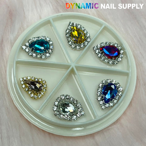 A heart-shaped nail supply wheel showcases rhinestones in blue, yellow, pink, amber, and clear tones, each framed by tiny silver crystals. The Dynamic Nail Supply brand name is elegantly displayed in the top right corner. These delightful nail charms are beautifully highlighted against a soft, textured pink background.