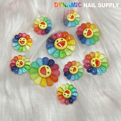 A collection of Smiling Face Flower / Happy Sunflower charms for nails from Dynamic Nail Supply is showcased against a plush, white fur backdrop. These lively accessories boast multicolored petals and joyful expressions, with the brand name "Dynamic Nail Supply" prominently printed in the top right corner.