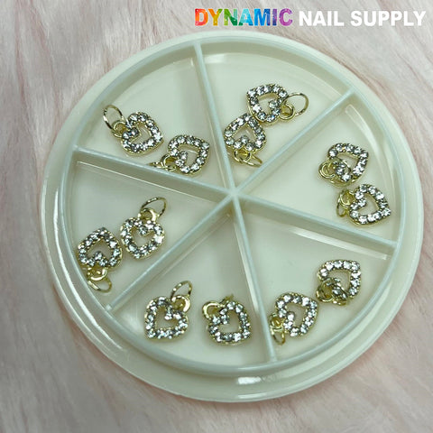 A circular container boasting six compartments displays the Heart-shaped Dangles Nail Charms, adorned with tiny rhinestones. The soft, light pink background accentuates the presentation. "Dynamic Nail Supply" is prominently featured in a gradient color at the top right corner, highlighting their exquisite nail accessories.