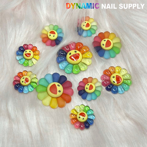 Flower-shaped nail art decorations and resin charms from Dynamic Nail Supply are arranged on a soft, white fur background. These "Smiling Face Flower" or "Happy Sunflower" charms feature petals in vibrant rainbow colors and a yellow center adorned with a cheerful, open-mouthed smiley face. The brand name, DYNAMIC NAIL SUPPLY, is displayed in the top right corner.