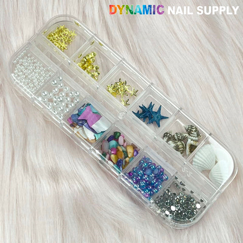 A transparent plastic organizer box features an assortment of nail art decorations, such as seashell charms, pearls, starfish sequins, and colorful rhinestones. Ideal for summer nail art designs, the compartments present various shapes and sizes against a soft pink fabric background. The top showcases Dynamic Nail Supply in style.