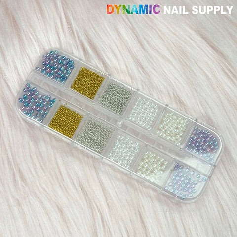 A clear plastic container with twelve compartments, filled with a colorful array of nail art beads including shimmering gold and silver round sphere balls and pearls, is placed on a textured, light-colored surface. The container holds beads in metallic finishes, white tones, and various pastel colors. Text in the top right corner reads Dynamic Nail Supply.