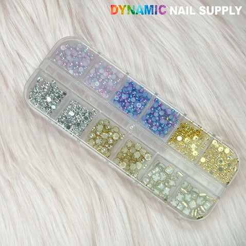 A White Pearl and gold silver ball beads set is placed on a soft, fluffy surface. It is organized in a 12-grid case containing assorted rhinestones and gold sphere balls in an array of colors like pink, blue, silver, and clear. The grids are neatly arranged in a transparent rectangular case featuring the brand name Dynamic Nail Supply printed in the top right corner.