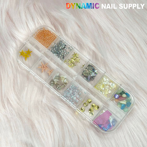 A clear rectangular organizer from Dynamic Nail Supply sits on a plush light pink surface, filled with summer nail art essentials like miniature beads, seashell charms, metallic studs, and colorful gemstones. In the top right corner, "DYNAMIC NAIL SUPPLY" is displayed in vibrant letters.