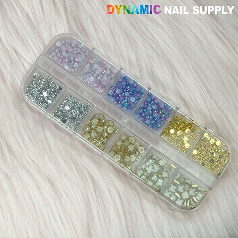 A clear plastic 12-grid case holds a variety of nail art rhinestones, including gold and silver sphere balls. The stones are sorted by color: silver, pink, blue, gold, and iridescent. Set against a soft beige fuzzy fabric backdrop, this White Pearl and Gold Silver Ball Beads Set from Dynamic Nail Supply is perfect for all your creative needs.