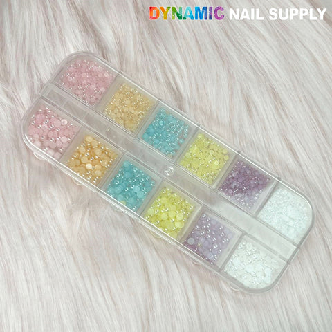 A set of twelve light pastel color pearls and half-sphere ball beads from Dynamic Nail Supply is arranged in a clear plastic organizer. The various pastel shades include pink, peach, orange, yellow, blue, purple, and white. The organizer is placed on a soft, fluffy surface with the brand name prominently displayed at the top right.