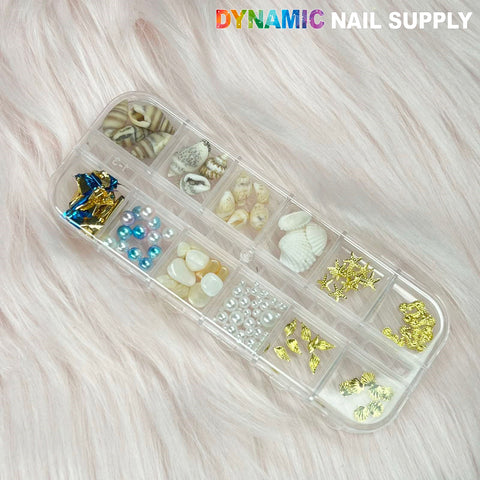A transparent nail art supplies organizer showcases the "Summer Nail Charm" collection, featuring seashell and starfish charms, gold accents, blue and white pearls, and golden embellishments. It rests on a light pink faux fur background with the text "DYNAMIC NAIL SUPPLY" displayed prominently in vibrant letters at the top.