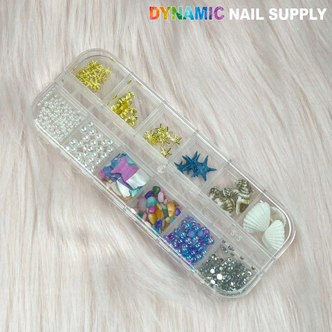 A divided container showcases a selection of summer-themed nail art decorations, featuring sequins like pearls, starfish, and seashell charms for nail design. The scene is set against a light, fluffy background with the brand name Dynamic Nail Supply displayed in the top right corner.