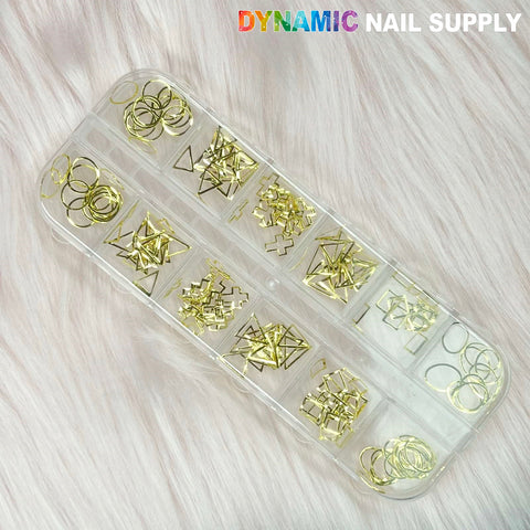 A transparent 12-grid box contains an assortment of Most Popular Nail Sequins and Rivet for Nails Designer set057 from Dynamic Nail Supply, featuring gold metallic geometric shapes such as circles, triangles, and stars. The organizer is placed on a cream-colored textured fur surface with the brand name Dynamic Nail Supply displayed in the top right corner.