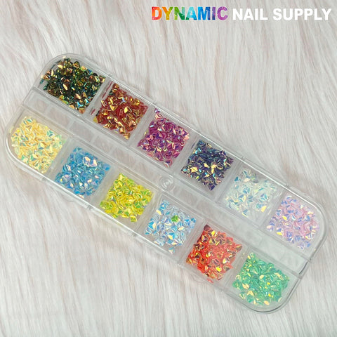 A plastic container holds twelve compartments, each packed with Colorful Iridescent Diamond-shaped glitters and vibrant nail art charms in hues such as green, yellow, blue, pink, and purple. The top right corner prominently features the Dynamic Nail Supply brand in colorful text against a backdrop of soft white fabric.