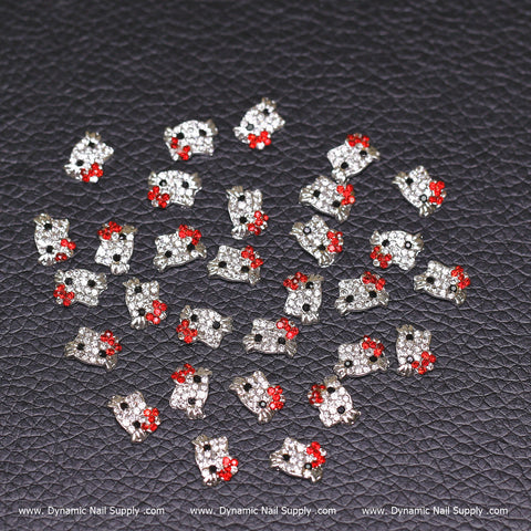 A set of 30 Metal Hell0 Kitty Design Nail Charms in red, made from zircon alloy, features small decorative embellishments shaped like skulls with silver and red rhinestone details. The charms are scattered artfully on a textured black surface, creating the perfect accessory for nail enthusiasts. The shiny finish highlights the Dynamic Nail Supply brand at www.DynamicNailSupply.com.