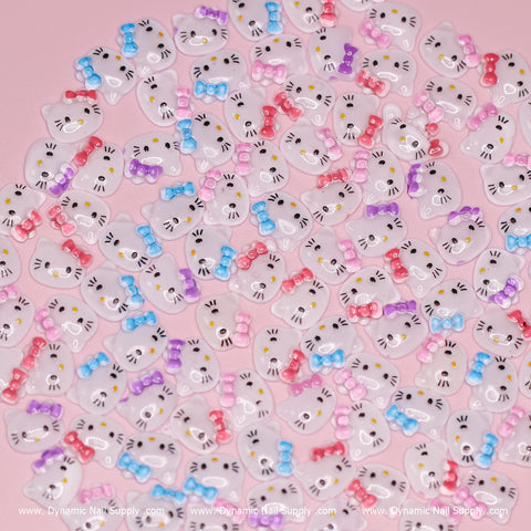 A delightful assortment of the "Mixed Colors" 100 pcs Cute Resin Hello Kitty Design Nail Charms from Dynamic Nail Supply is displayed against a pink background. These resin nail charms showcase white cat faces adorned with bows in vibrant hues of red, blue, pink, and purple. Each charm features a tiny gold cat nose and black whiskers, forming a playful and whimsical design.