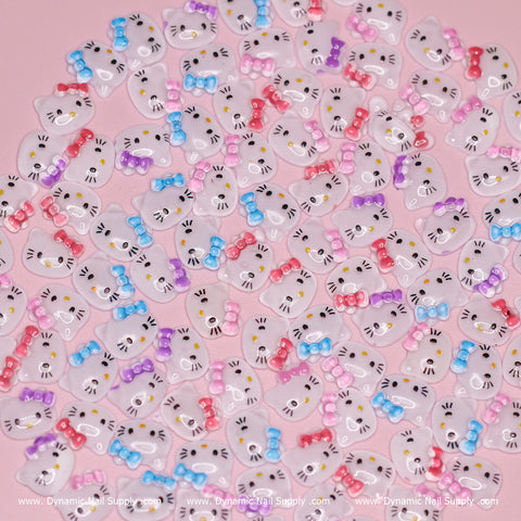 The (Mixed Colors) 100 pcs Cute Resin Hell0 Kitty Design Nail Charms by Dynamic Nail Supply feature small, decorative resin charms shaped like white cat faces with colorful bows—pink, red, blue, and purple—on a pink background. Each charm is adorned with painted whiskers, eyes, and a nose in a cartoon style reminiscent of Hello Kitty, making them perfect for your dynamic nail supply kit!