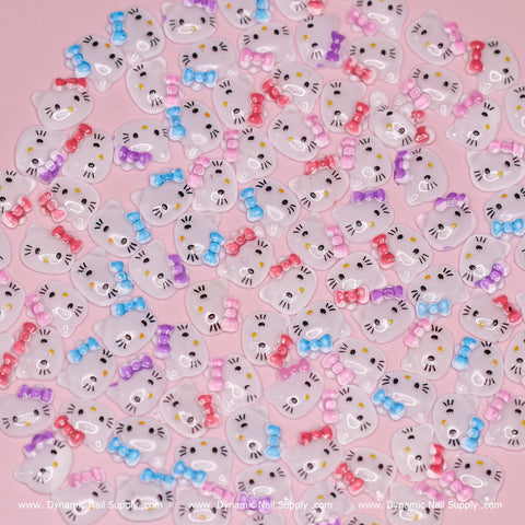 A collection of adorable nail art decorations, featuring cartoon cat faces with whiskers and colorful bows akin to Hello Kitty, embellishes a soft pink backdrop. These resin nail charms from Dynamic Nail Supply, available in mixed colors including white adorned with pink, blue, purple, or red bows, prominently display the brand's repeated logos at the bottom.