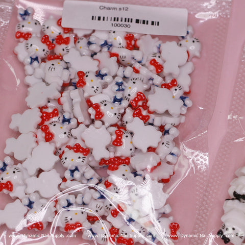 A detailed look at the opened packaging showcases adorable resin nail charms in the shape of a white cat with a red bow and blue overalls, reminiscent of Hello Kitty. The label indicates it is Charm s12 with the number 100030. Positioned against a pink background, the package displays seams and includes the website www.DynamicNailSupply.com from Dynamic Nail Supply.