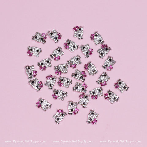 Tiny magenta bow-shaped zircon alloy nail charms inspired by Hello Kitty are scattered over a pink surface. Each charm sparkles with clear and pink stones, reflecting the essence of Hello Kitty design. The background displays the "Dynamic Nail Supply" text repeatedly along the lower edge.