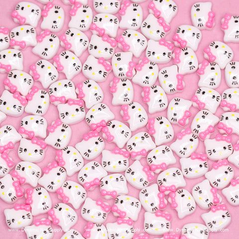 An assortment of small, glossy nail charms featuring Hello Kitty designs with pink bows and yellow noses is beautifully displayed across a pink surface. The cat faces are adorned with black eyes, whiskers, and mouths. This playful setup by Dynamic Nail Supply brings a whimsical pattern to life, perfect for their Cute Resin Hell0 Kitty Design Nail Charms in Pink.