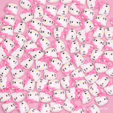 A pile of small, white cute resin Hello Kitty design nail charms with pink bows is scattered on a pink surface. The cats feature yellow noses, black whiskers, and black eyes. Each face is uniformly styled like a cartoon character—perfectly cute for Dynamic Nail Supply’s (Pink) 100 pcs collection.