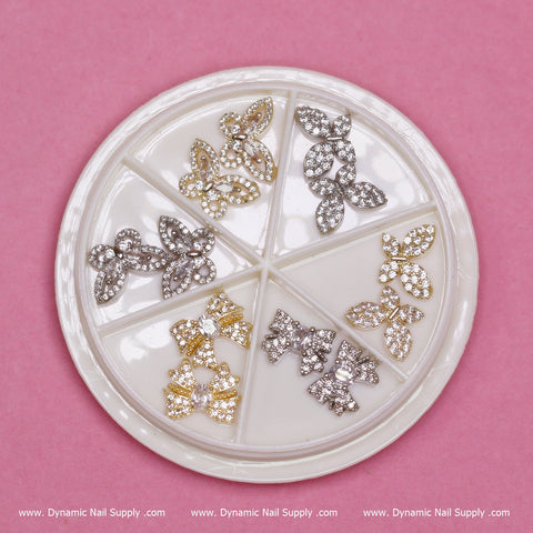 A circular nail art decoration wheel set against a pink background showcases six compartments filled with diverse rhinestone designs, including the Dynamic Nail Supply's (Gold) 12 pcs Unique Butterfly Rhinestone Charms for Nails Art Designer and floral shapes in silver and gold tones. The brand name "Dynamic Nail Supply" is elegantly repeated along the bottom edge.