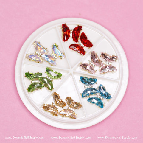 A stunning palette by Dynamic Nail Supply, featuring their (Iridescent 04) Butterfly Rhinestone Charms for Nails Art Designers, is beautifully showcased on a pink background. The set includes twelve pieces of colorful marquise-shaped rhinestones in shades of green, opalescent white, yellow, red, pink, blue, and purple—ideal for enhancing any creative nail design.