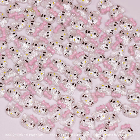 A collection of adorable resin nail charms is spread across a pink surface. Each charm showcases a Hello Kitty design with glittery silver faces, black eyes, pink bows, and yellow nose details. The manufacturer’s name displayed at the bottom is Dynamic Nail Supply.