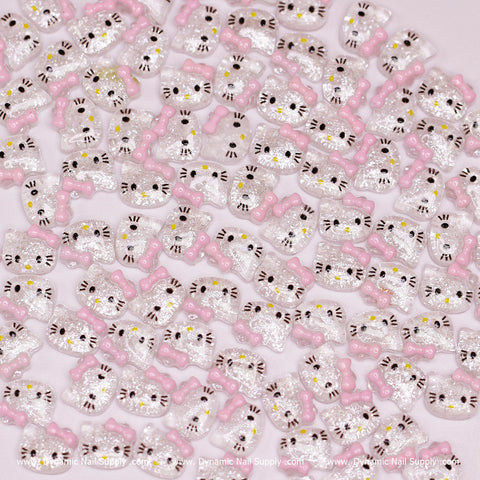 A collection of the (Clear & Pink) 100 pcs Cute Resin Hell0 Kitty Design Nail Charms inspired by Hello Kitty features shiny resin charms with cartoon cat motifs, showcasing pink bows and white faces. Each charm is detailed with black eyes and nose, artfully arranged on a light pink surface. The backdrop subtly displays the brand name: Dynamic Nail Supply.