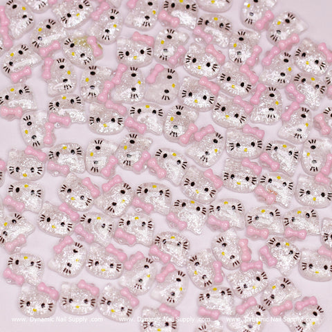 A set of charming resin nail embellishments shaped like Hello Kitty faces, featuring a delightful pink bow and glittery accents, with black whiskers and eye details. These charms are scattered on a light pink background, making them perfect for any nail art design. The Clear & Pink 100 pcs Cute Resin Hell0 Kitty Design Nail Charms are offered by Dynamic Nail Supply.