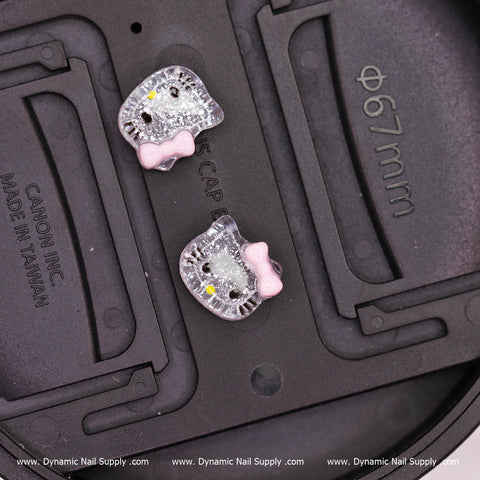 Close-up of two small, adorable resin nail charms shaped like cartoon cat faces with pink bows. They have shiny rhinestone accents and black outlines. The Hell0 Kitty Design Nail Charms from Dynamic Nail Supply are set against a black circular background that includes measurements and manufacturing details.