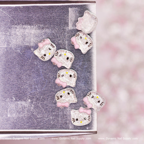 The image features a selection from Dynamic Nail Supply's (Clear & Pink) 100 pcs Cute Resin Hell0 Kitty Design Nail Charms, arranged in a diagonal line. These charms have glittery silver surfaces adorned with pink bows and yellow accents. The backdrop is metallic with a textured appearance, and the Dynamic Nail Supply logo is subtly visible.