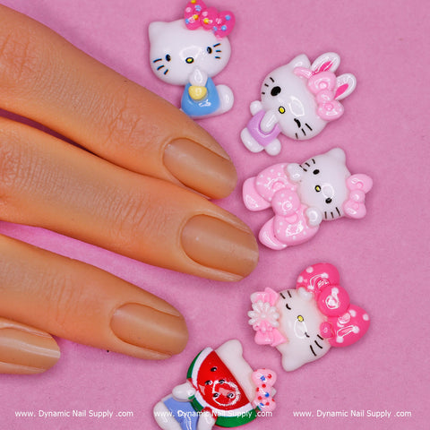 A hand with a nude manicure rests on a pink surface beside the charming 10 pcs Hello Kitty Charm set from Dynamic Nail Supply. These adorable nail charms display Hello Kitty in various poses, holding fun items like an ice cream cone and a watermelon slice, all adorned with pink bows and flowers—making them the perfect accent for creative nail art designs.