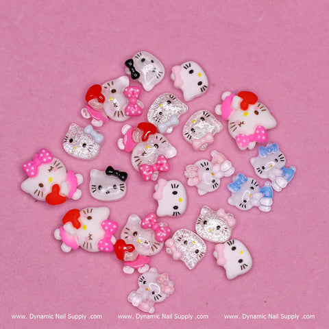 A delightful set of cat-themed nail charms, echoing the charm of Hello Kitty, presented on a pink background. This 20-piece collection by Dynamic Nail Supply features an assortment of designs with bows and facial expressions in shades of white, pink, and red. Each charm is a stylized cat character with unique and playful features.