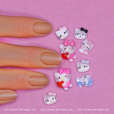 A hand with a neutral nail polish color is displayed against a soft pink background. Nearby, twenty colorful Hello Kitty Charms present delightful nail art designs, including bows and hearts. The decorations are neatly arranged in a cluster, offering inspiration from Dynamic Nail Supply's 20 pcs Random Hello Kitty Charm set (Mixed shapes) for Nail Art Design.